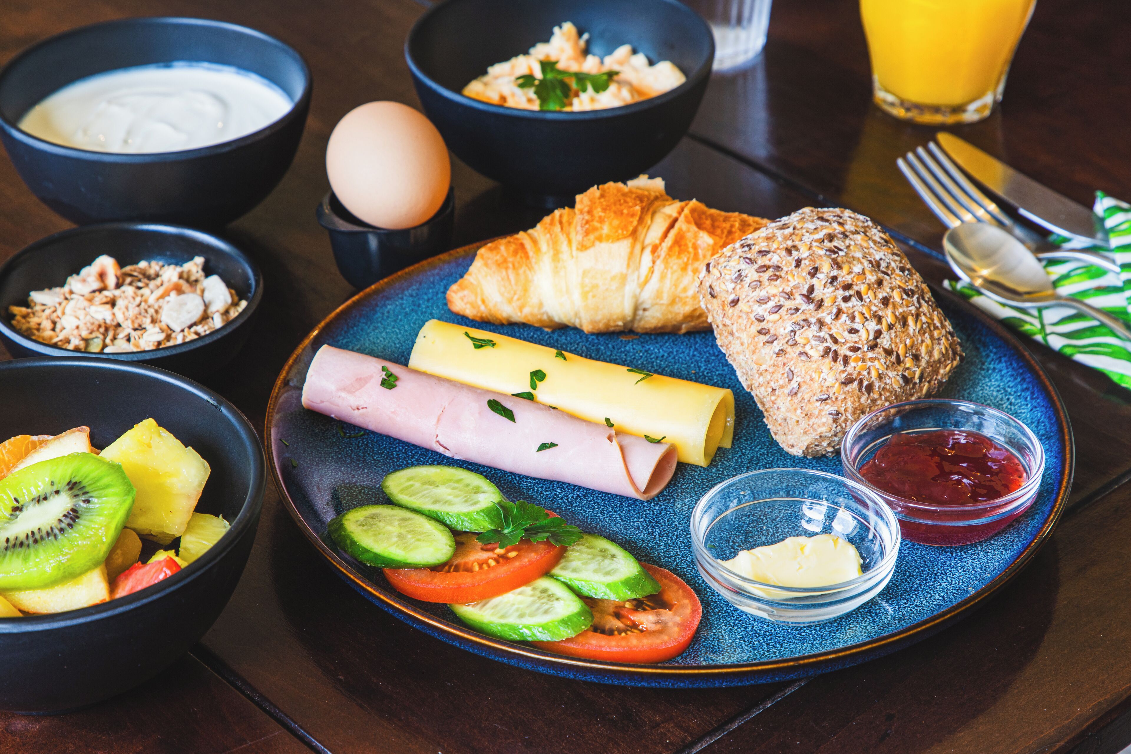 Een ontbijttafel met een bord eten, inclusief croissant, kaas, ham en groenten, en schaaltjes yoghurt, granola en fruit.