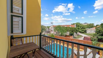 Varanda com mesa e cadeiras de madeira, vista para uma piscina e prédios com árvores.