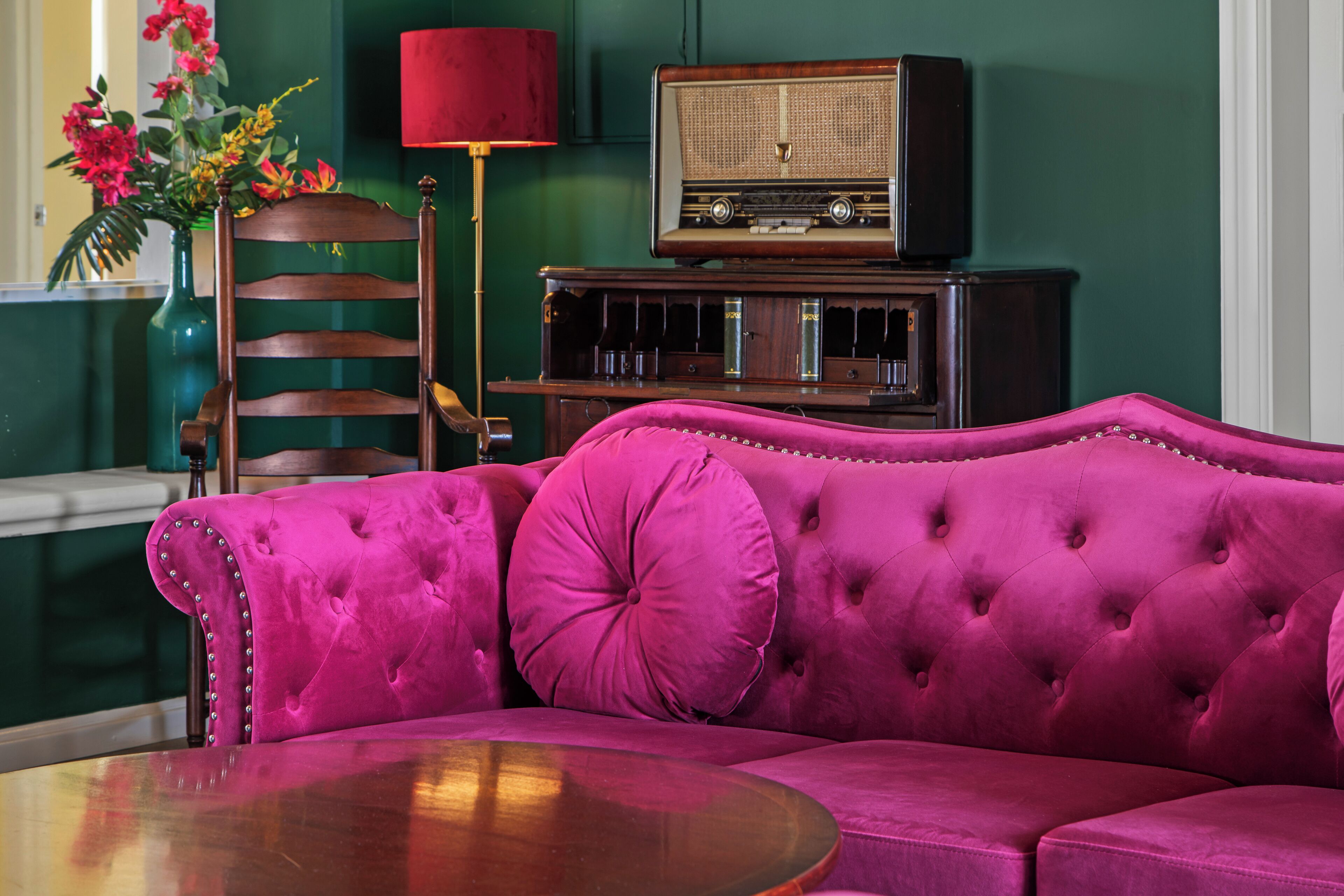 Una habitación con un sofá rosa con capitoné, una mesita de madera, una radio antigua y un jarrón con flores.