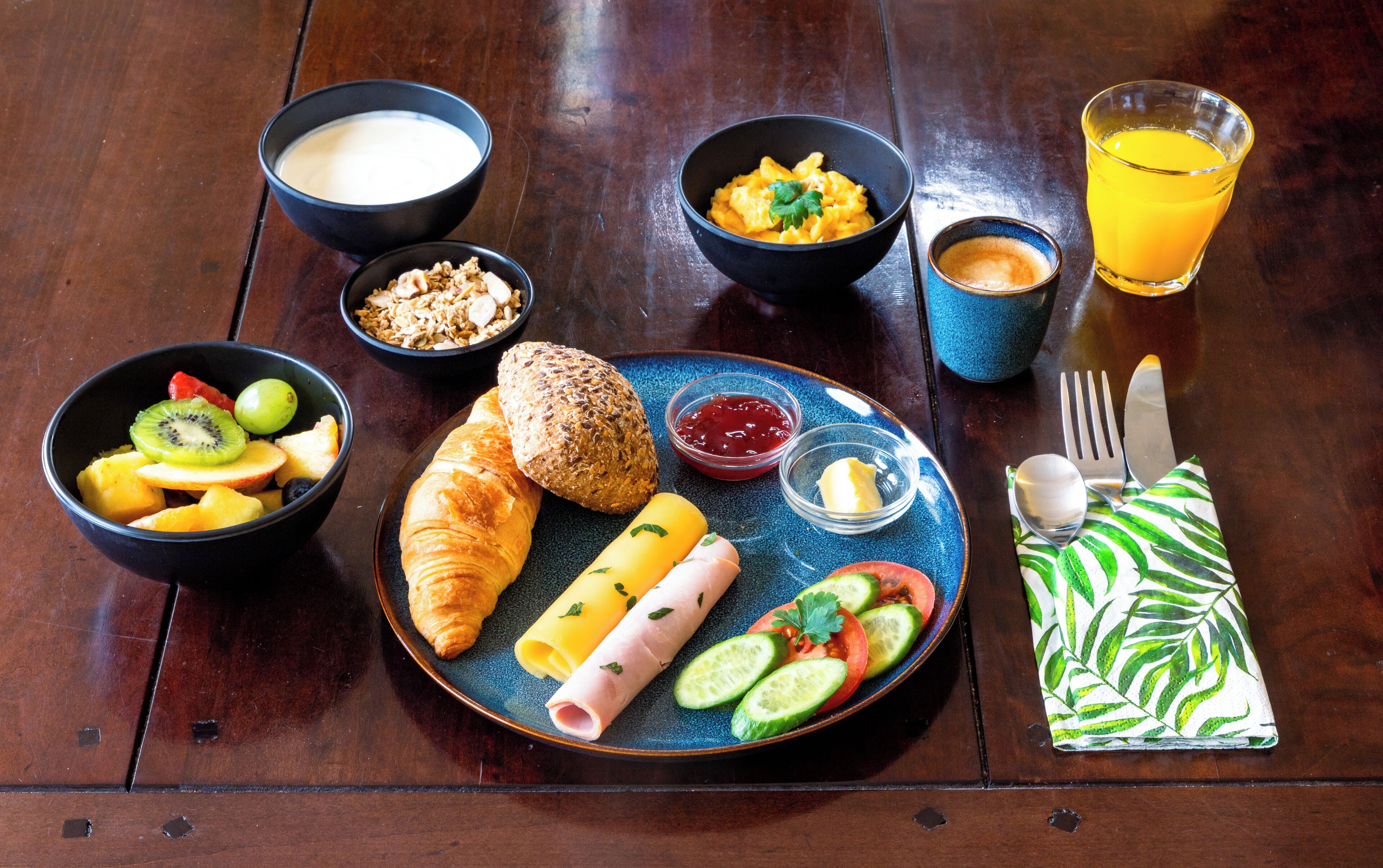 A breakfast setting with croissants, fruits, a plate of ham and cheese, bowls of yogurt and granola, a glass of orange juice, and a cup of coffee.