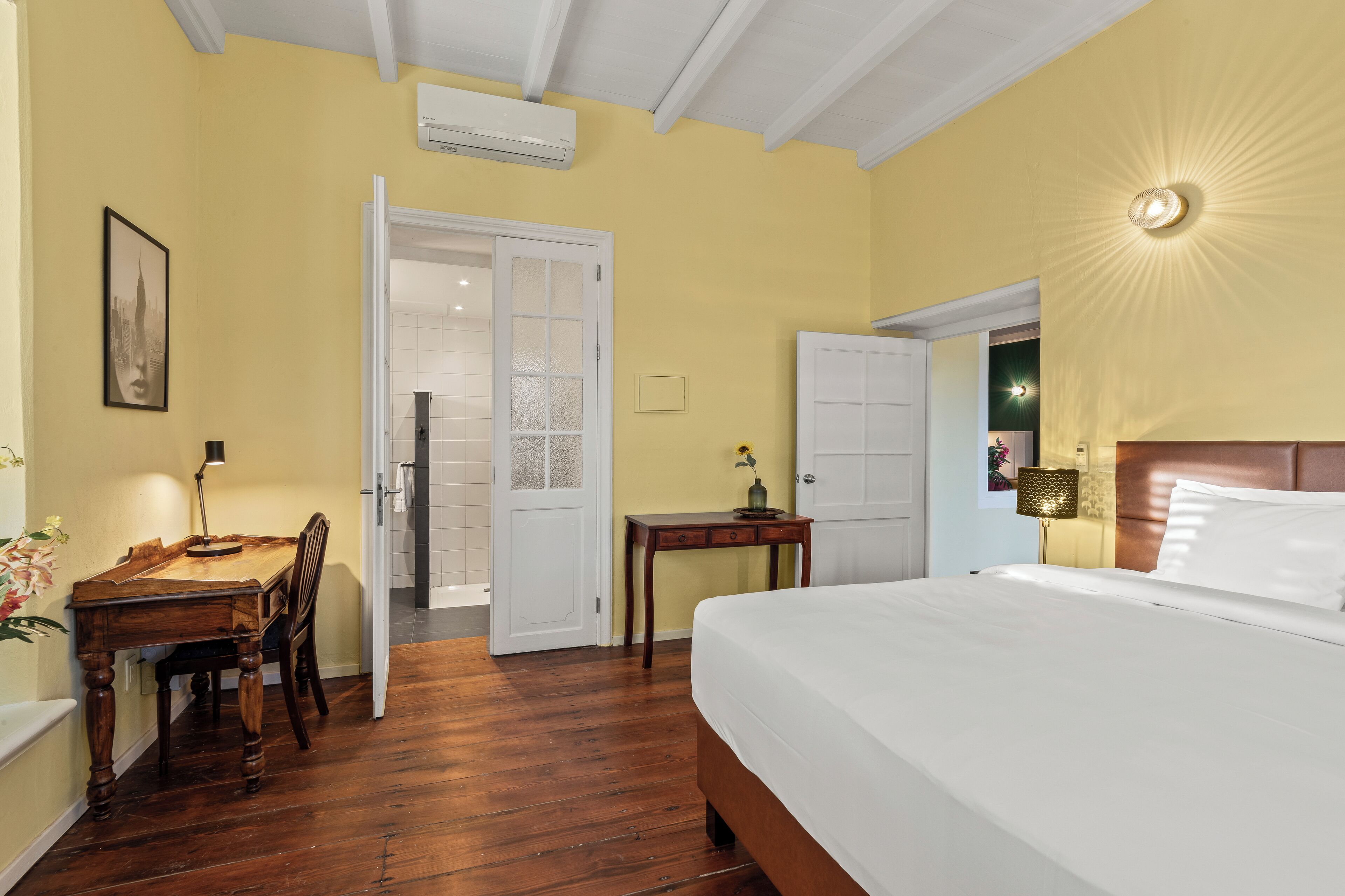 Ein Hotelzimmer mit Holzboden, einem Bett mit weißer Bettwäsche, einem Schreibtisch mit Stuhl und einem kleinen Tisch mit Lampe.