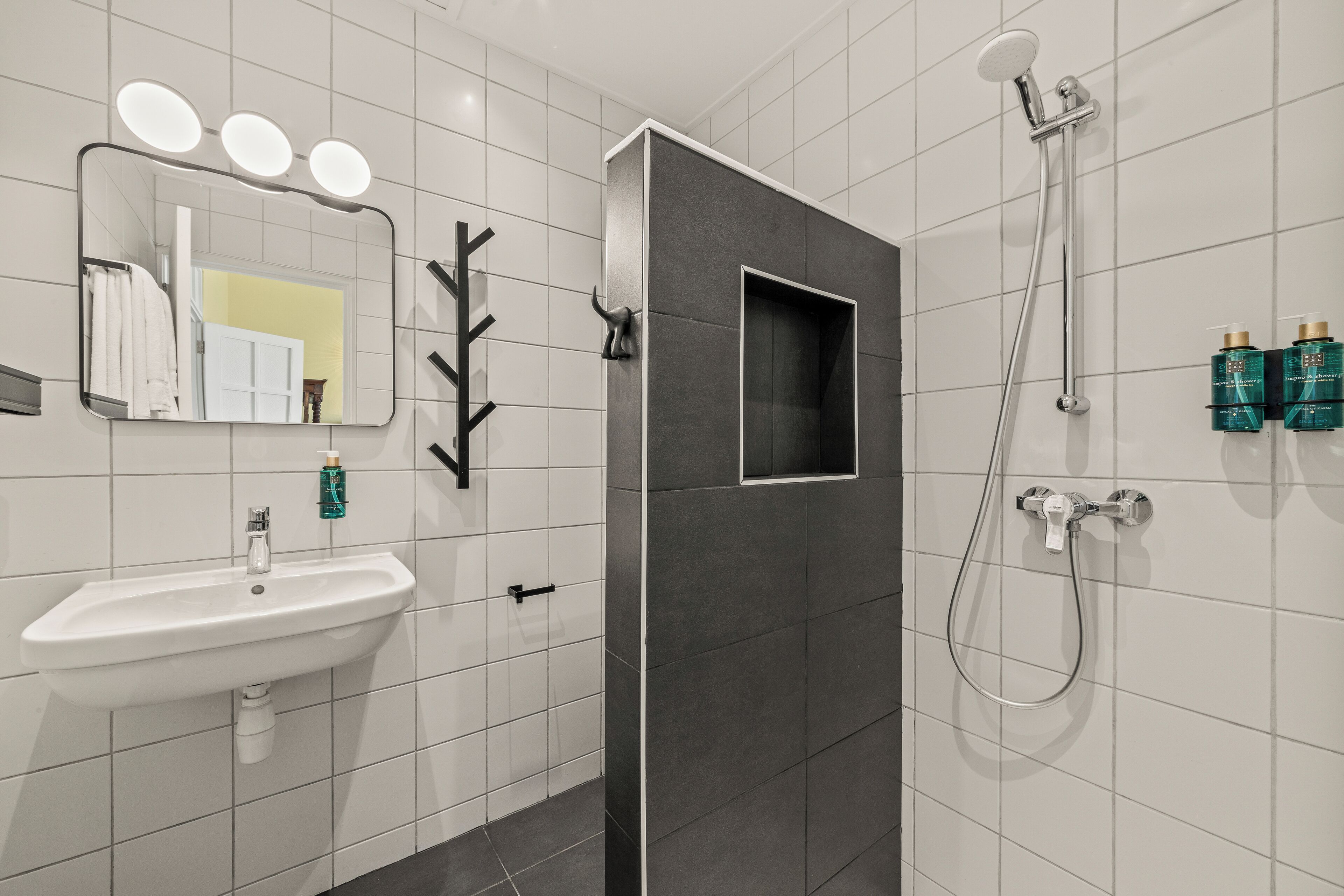 A modern bathroom with a white sink, a mirror with lights, a shower, and toiletries on the wall.