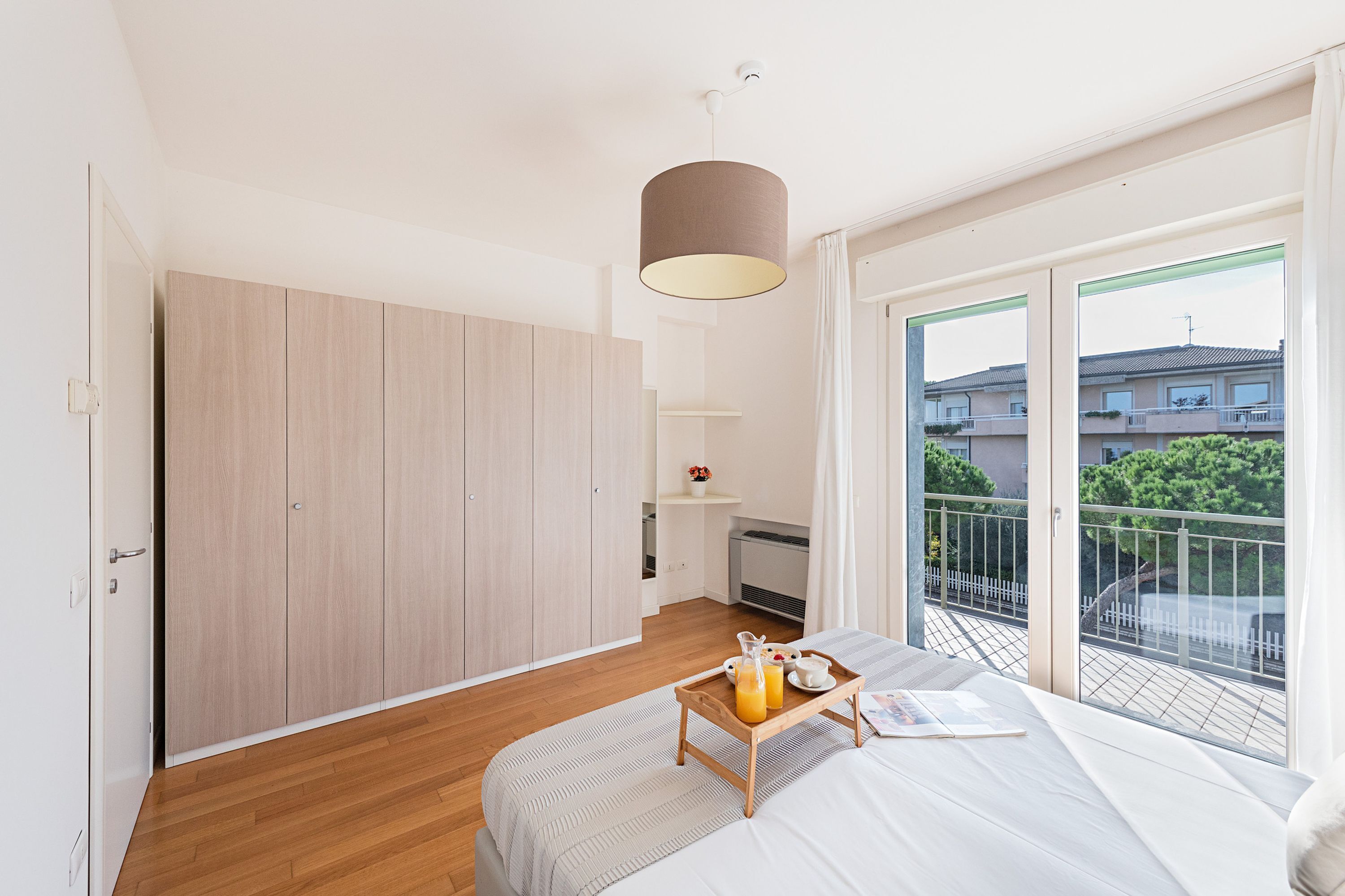 Een slaapkamer met een bed, een nachtkastje met een ontbijtje erop, een kledingkast en een balkon met uitzicht op gebouwen en groen.
