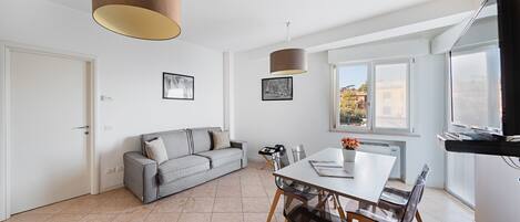 A modern living room with a grey sofa, a dining table with chairs, a TV, and a large window with a city view.