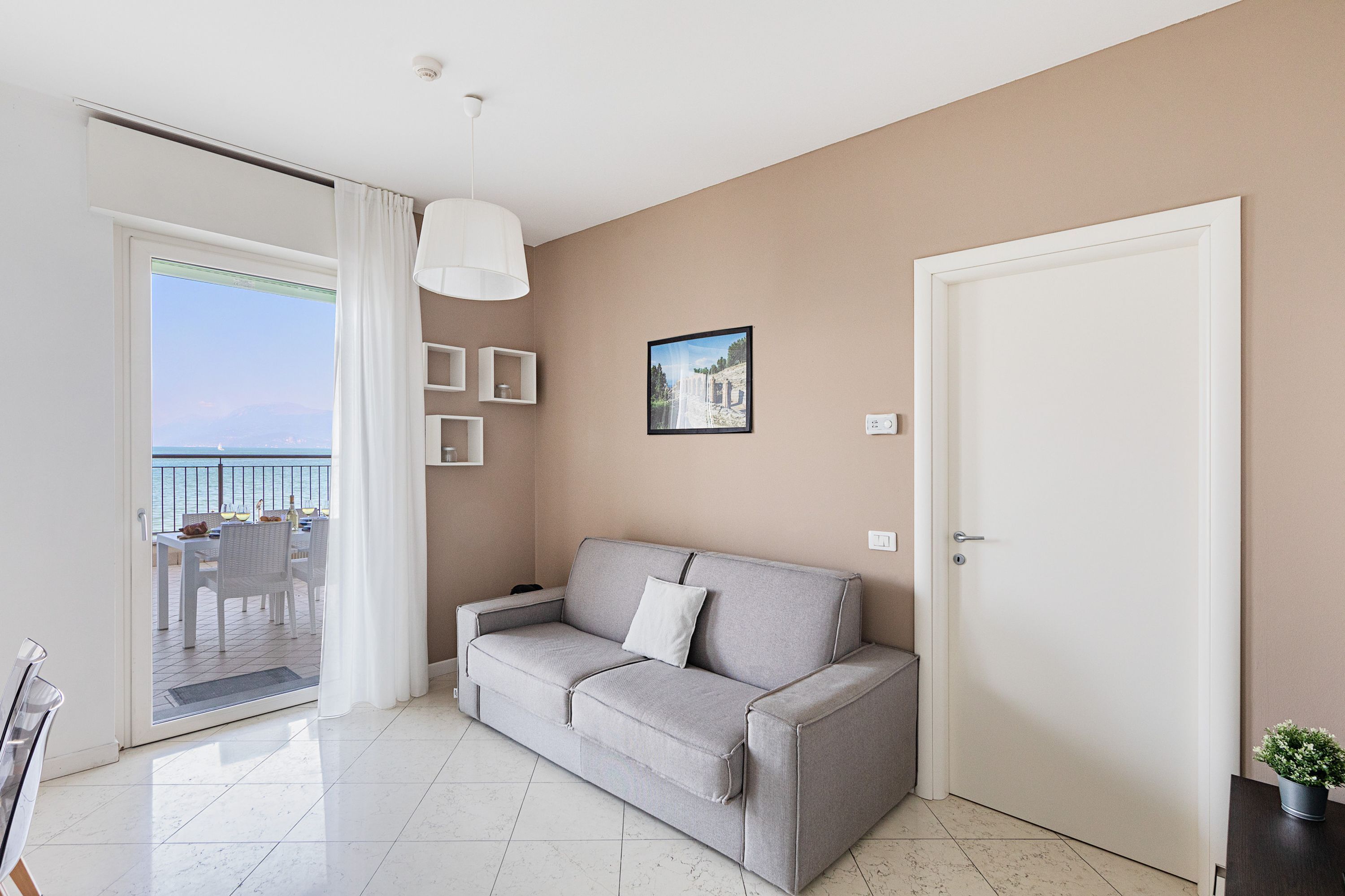 A modern living room with a grey sofa, a white door, and a view of the sea through a sliding glass door.