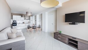 A modern living room with a sofa, dining area, kitchen, and a mounted TV. - Residenza Miralago - 24 Clodia - Locazione BK (Desenzano del Garda)