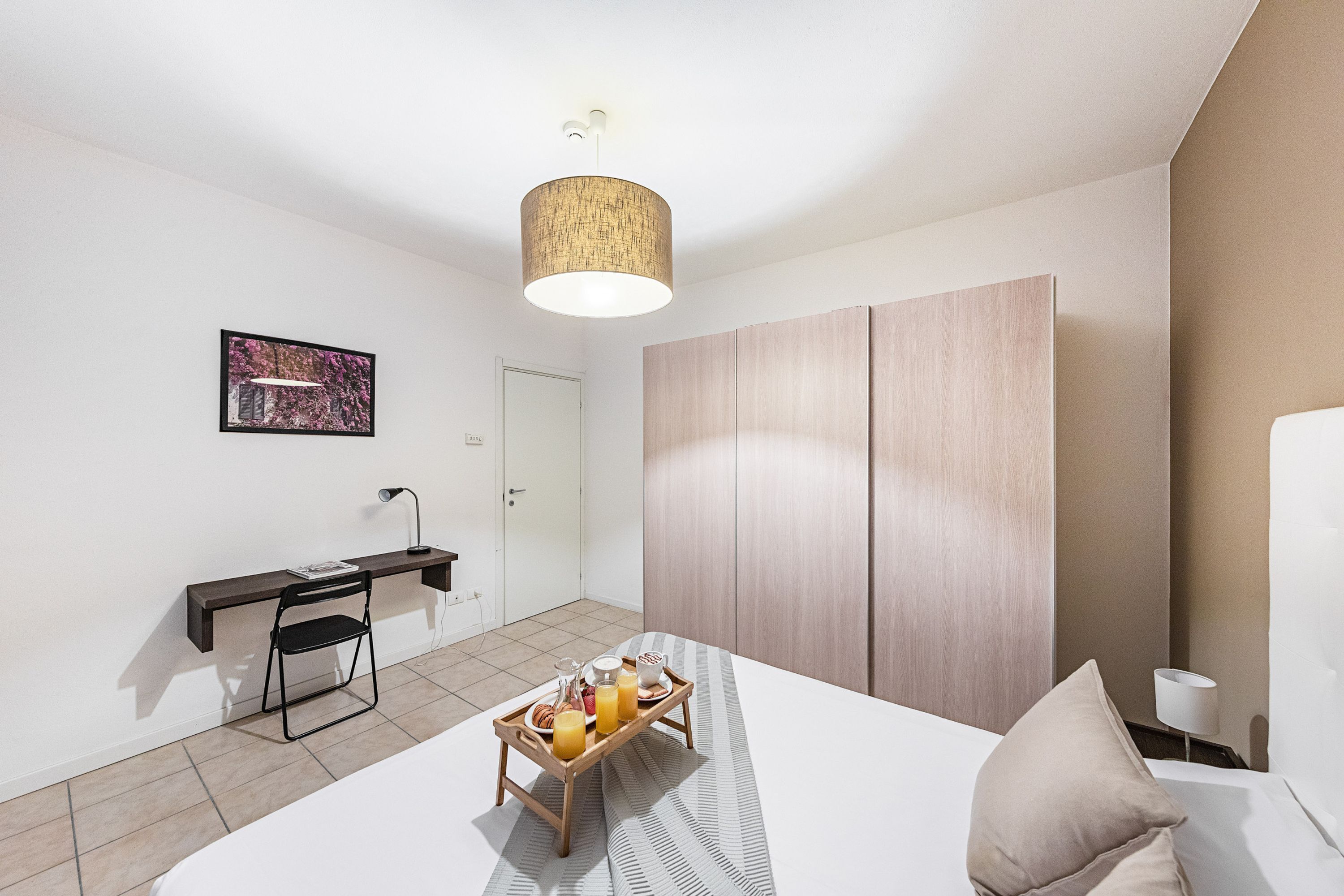 A modern hotel room with a bed, a desk with a chair, a tray with breakfast items, and a wardrobe.