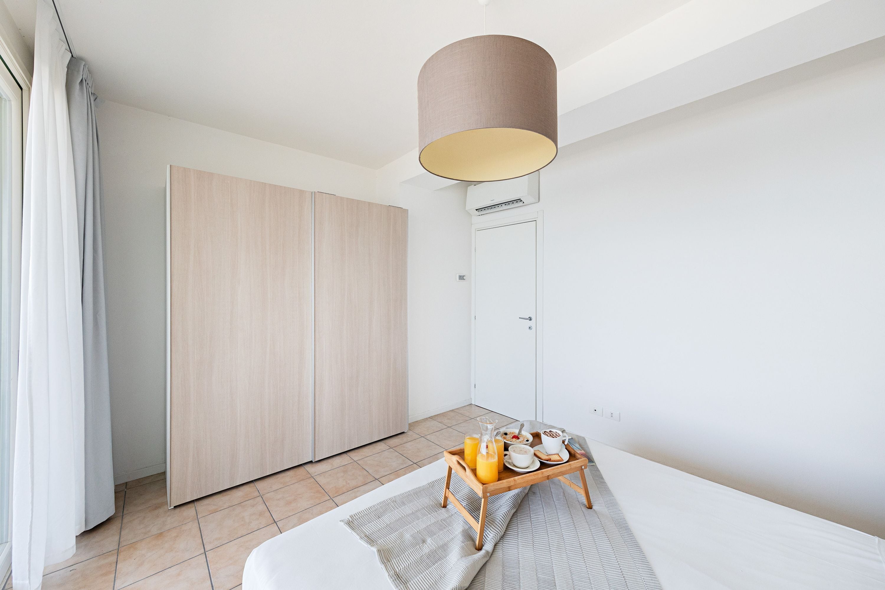 A modern bedroom with a wooden wardrobe, a bedside table with breakfast items, and a large window with curtains.