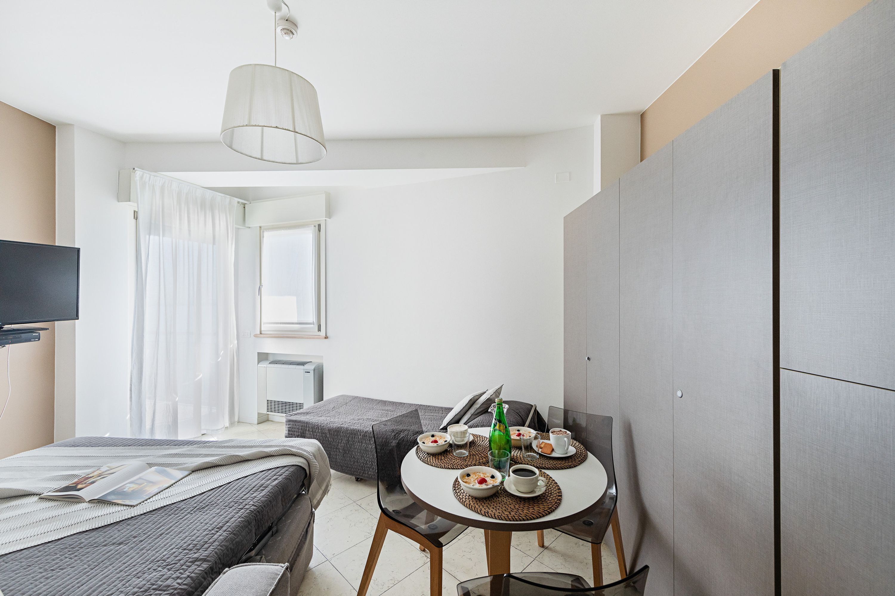 A modern hotel room with a bed, a dining table with breakfast, a TV, and a large wardrobe.