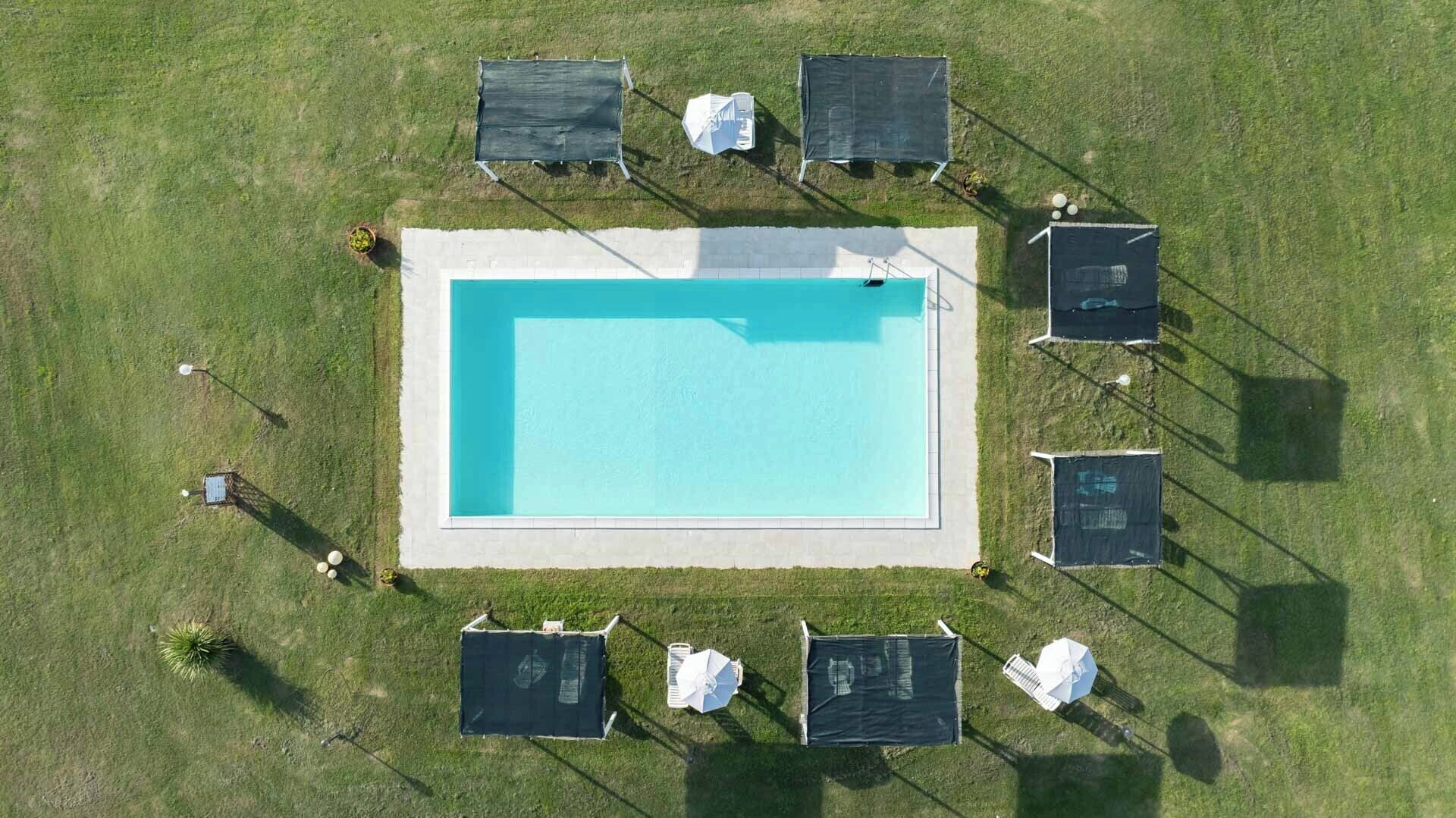 Une vue aérienne d’une piscine rectangulaire entourée d’une zone herbeuse comprenant plusieurs espaces de repos à l’ombre.
