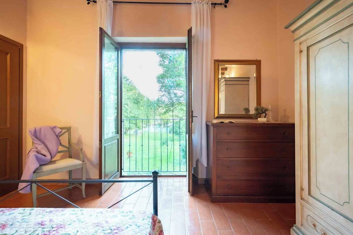 A bedroom with a bed, a dresser, a mirror, and a view of a green garden through an open door.