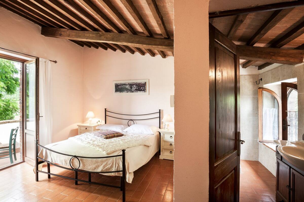 A bedroom with a bed, bedside tables, a window, and a bathroom visible through an open door.