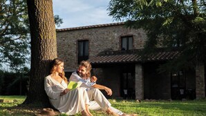 Dos personas sentadas sobre la hierba bajo un árbol, leyendo un libro, con un edificio de piedra al fondo.