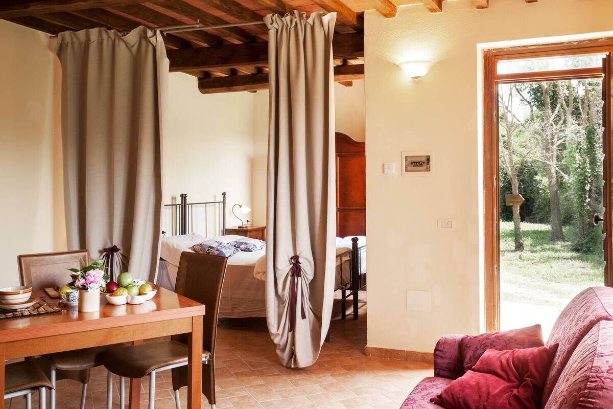 A cozy room with a wooden dining table, chairs, a bed with a headboard, and a view of the outdoors through a glass door.