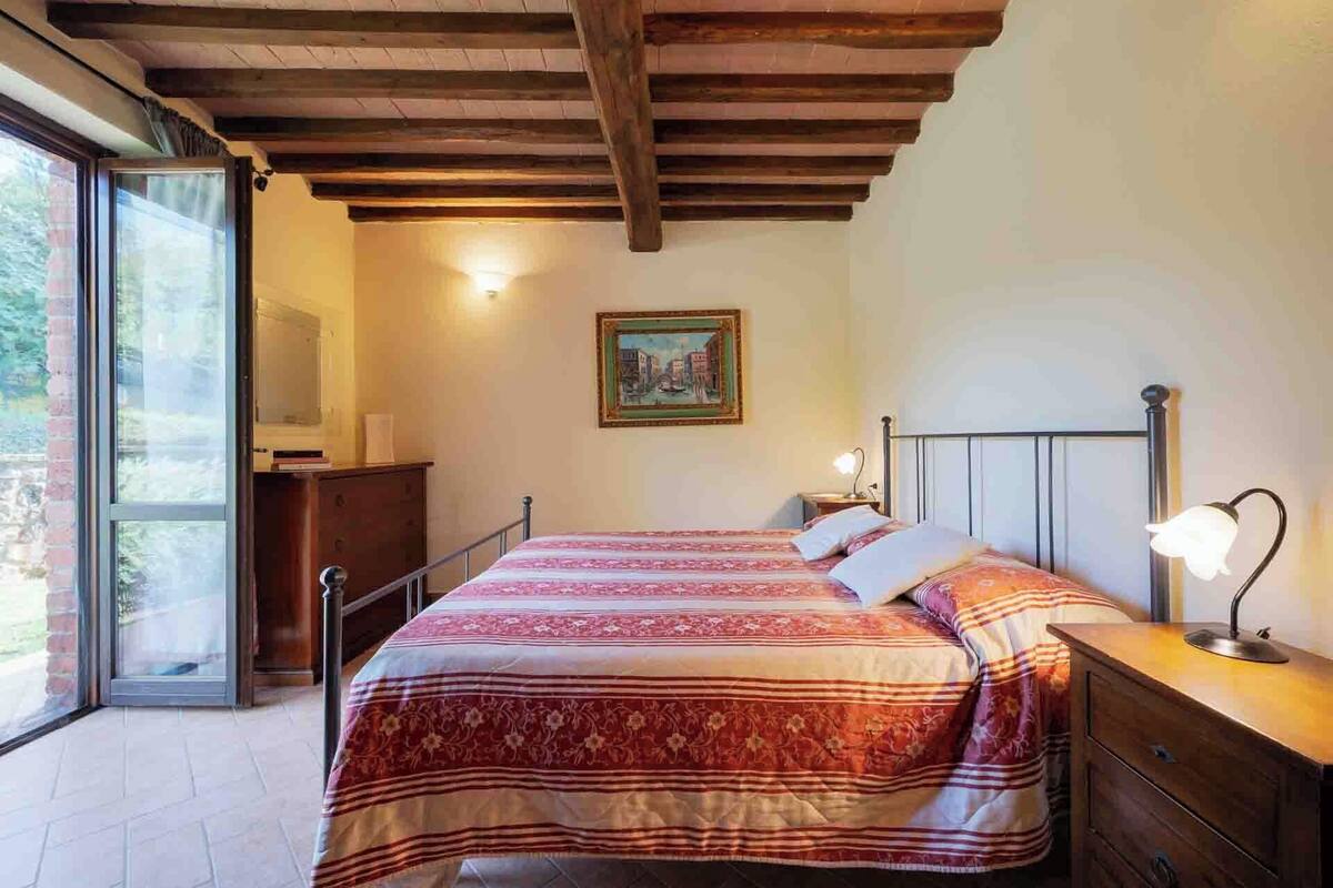 A bedroom with a bed, bedside tables, a nightstand, a painting on the wall, and a window with curtains.