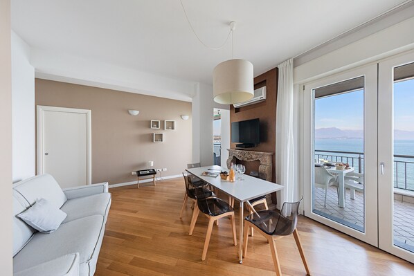 Una sala de estar moderna con sofá, mesa de comedor, sillas y televisor. Posee un balcón con vista al mar.