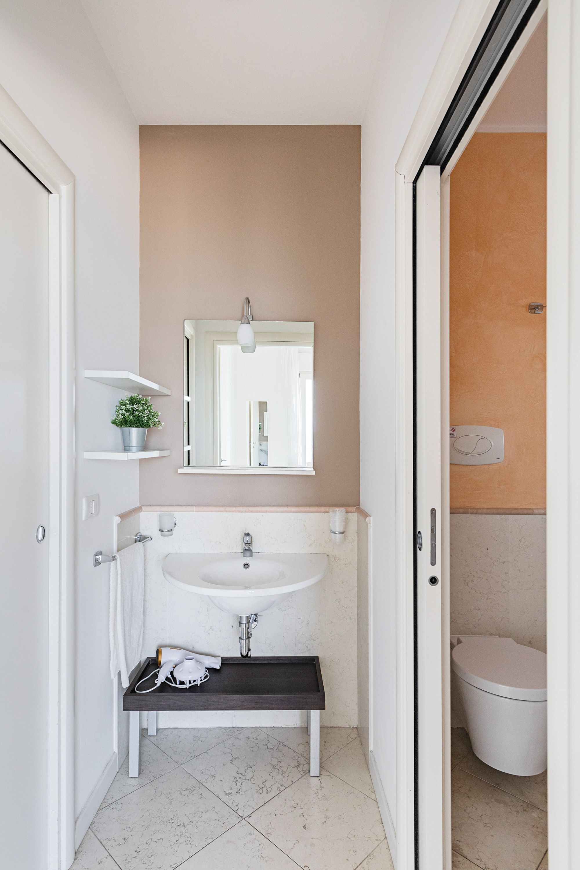 A bathroom with a white sink, mirror, and a toilet. There is a small shelf with a potted plant and a hairdryer on the counter.