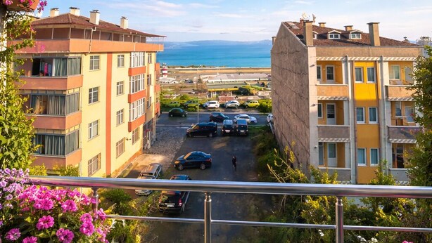 Un balcone con vasi di fiori, vista su una strada con auto parcheggiate e palazzi con balconi.