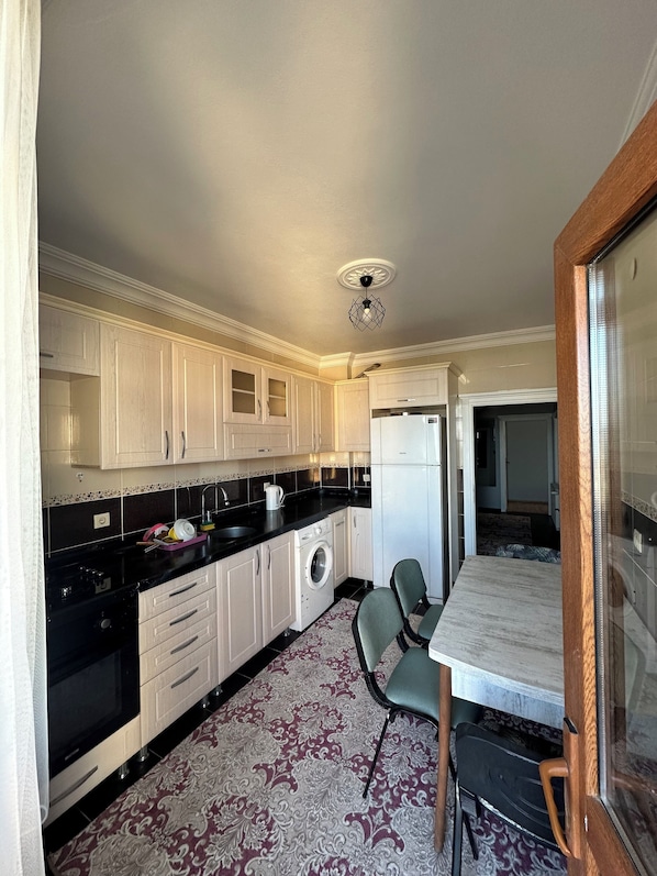 A compact kitchen with white cabinets, a black countertop, a washing machine, and a dining area with a table and chairs. - Kaya Rezidans (Trabzon)