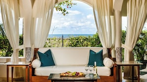 A cozy outdoor seating area with a white sofa, teal cushions, and a wooden coffee table. A bottle of wine and glasses are on the table, and a fruit platter is in front. The space is framed by white curtains and features a view of the ocean and greenery.