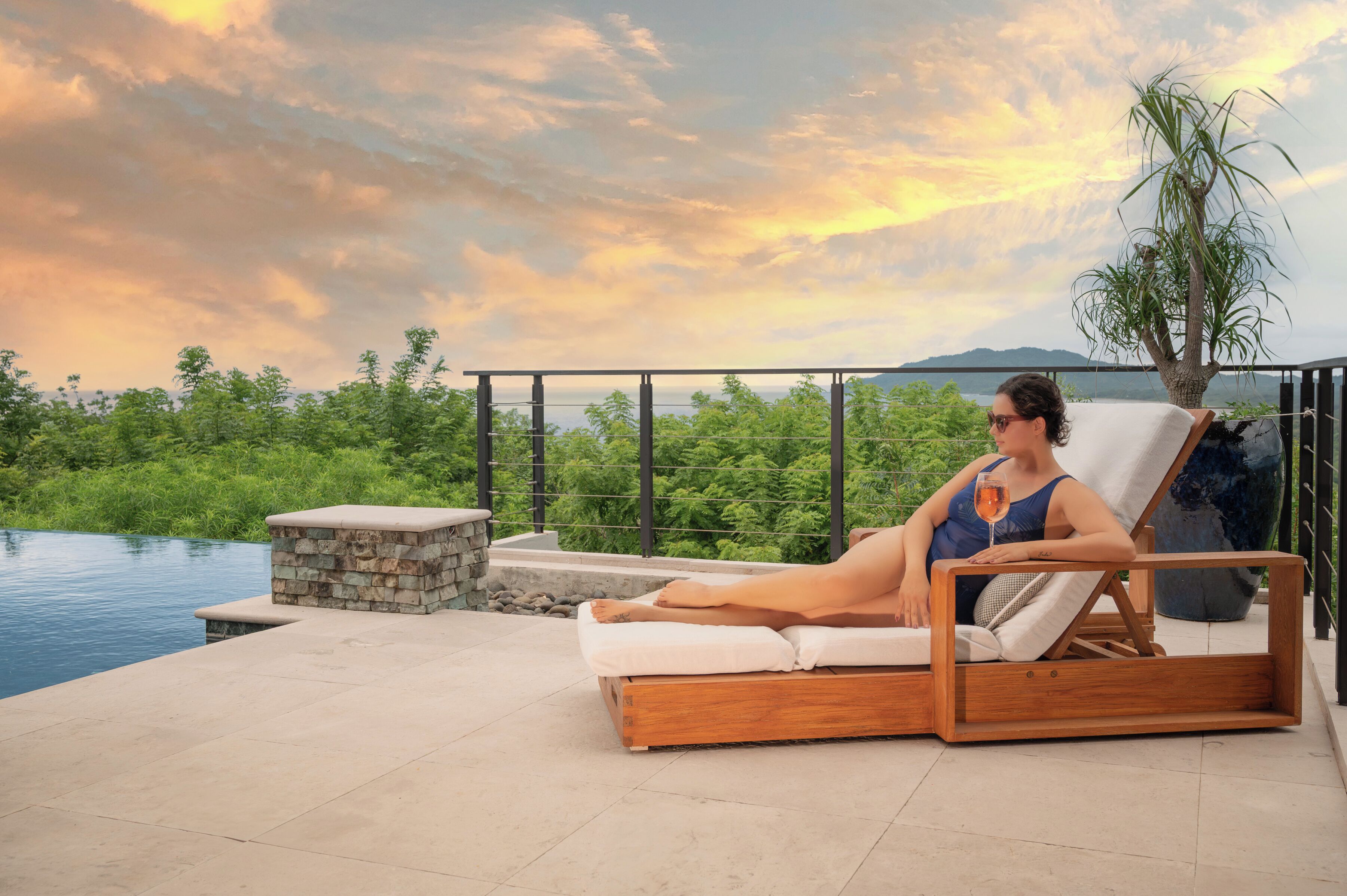 A person relaxing on a wooden lounge chair by a pool with a stone bench and a view of greenery and mountains.