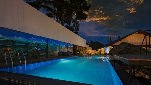 A swimming pool with a mural on the wall, surrounded by palm trees and a sunset sky. - Holidays Garden Resort (Kalpetta)