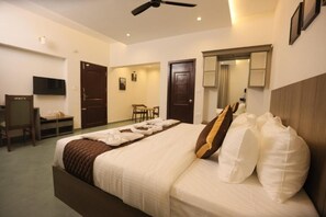 A hotel room with a bed, a desk, a chair, a television, and a ceiling fan. - Holidays Garden Resort (Kalpetta)