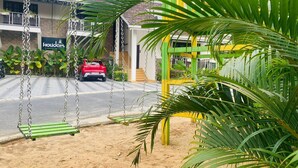 A hotel entrance with a swing set, a red car parked in front, and a sign that reads "Holidays Cardboard Services". - Holidays Garden Resort (Kalpetta)