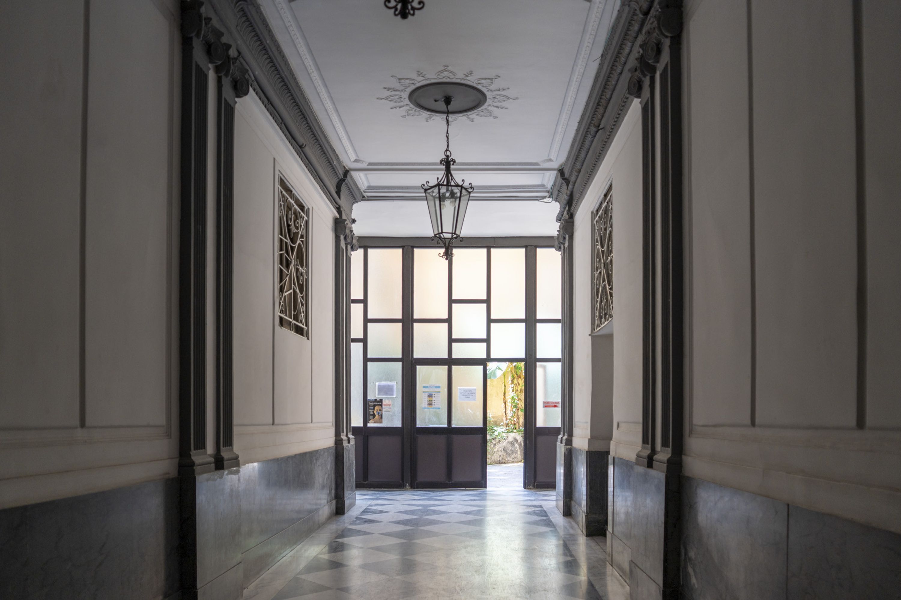 A hallway with a checkered floor, a chandelier, and a large window at the end.