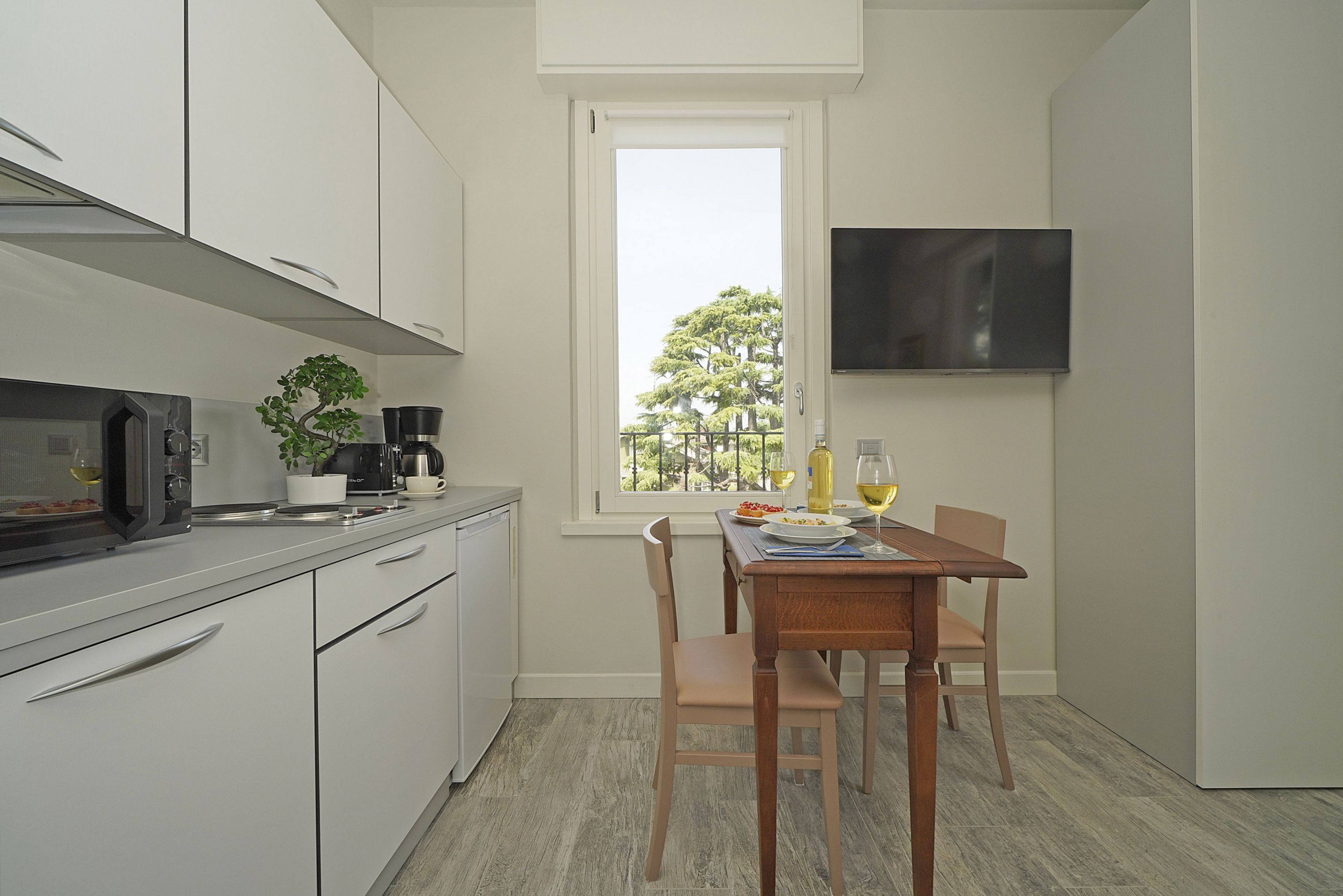 Une cuisine compacte avec des meubles blancs, un four à micro-ondes, une machine à café et une table à manger avec des chaises. Une fenêtre offre une vue sur les arbres à l’extérieur.