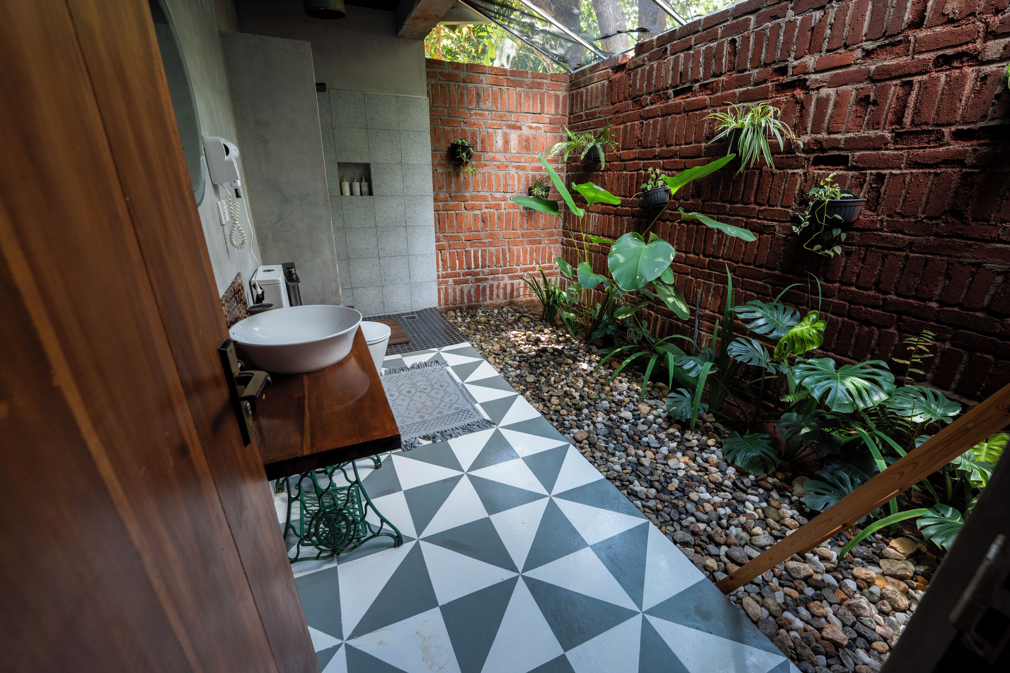 Un baño con lavamanos blanco, piso con estampado geométrico y pared de ladrillo con plantas en macetas.