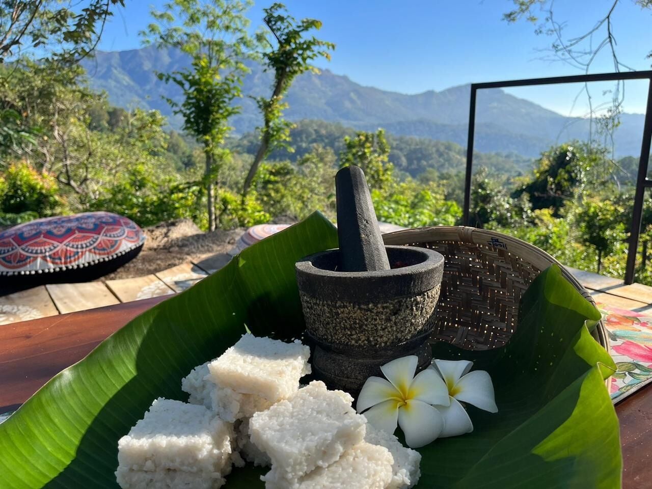 A traditional food setup with a mortar and pestle, rice, and a flower on a banana leaf, set against a mountainous backdrop.