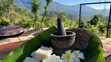 A traditional food setup with a mortar and pestle, rice, and a flower on a banana leaf, set against a mountainous backdrop.