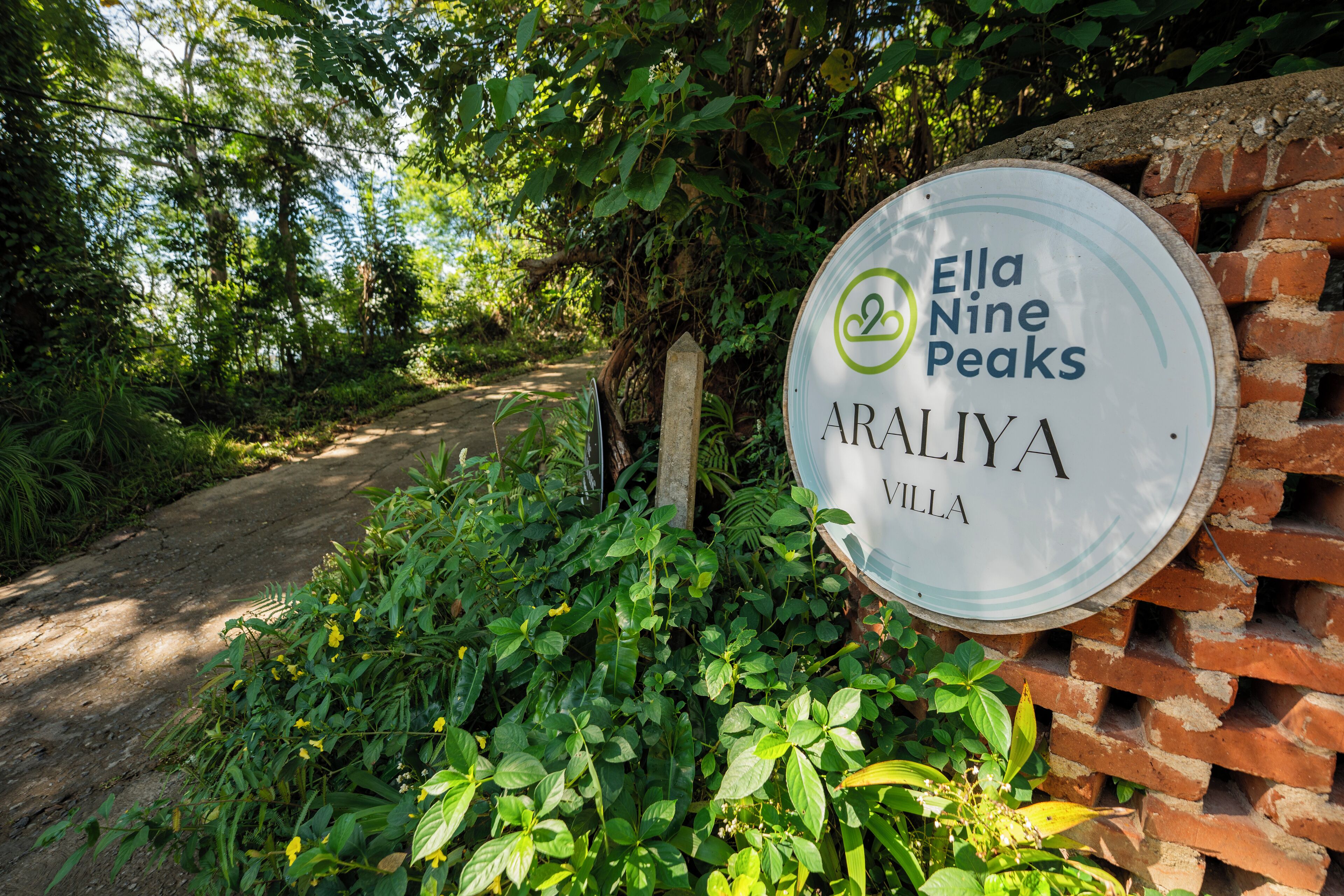Une enseigne indiquant « Ella Nine Peaks Araliya Villa », avec un mur de briques rustiques et une végétation luxuriante en arrière-plan.