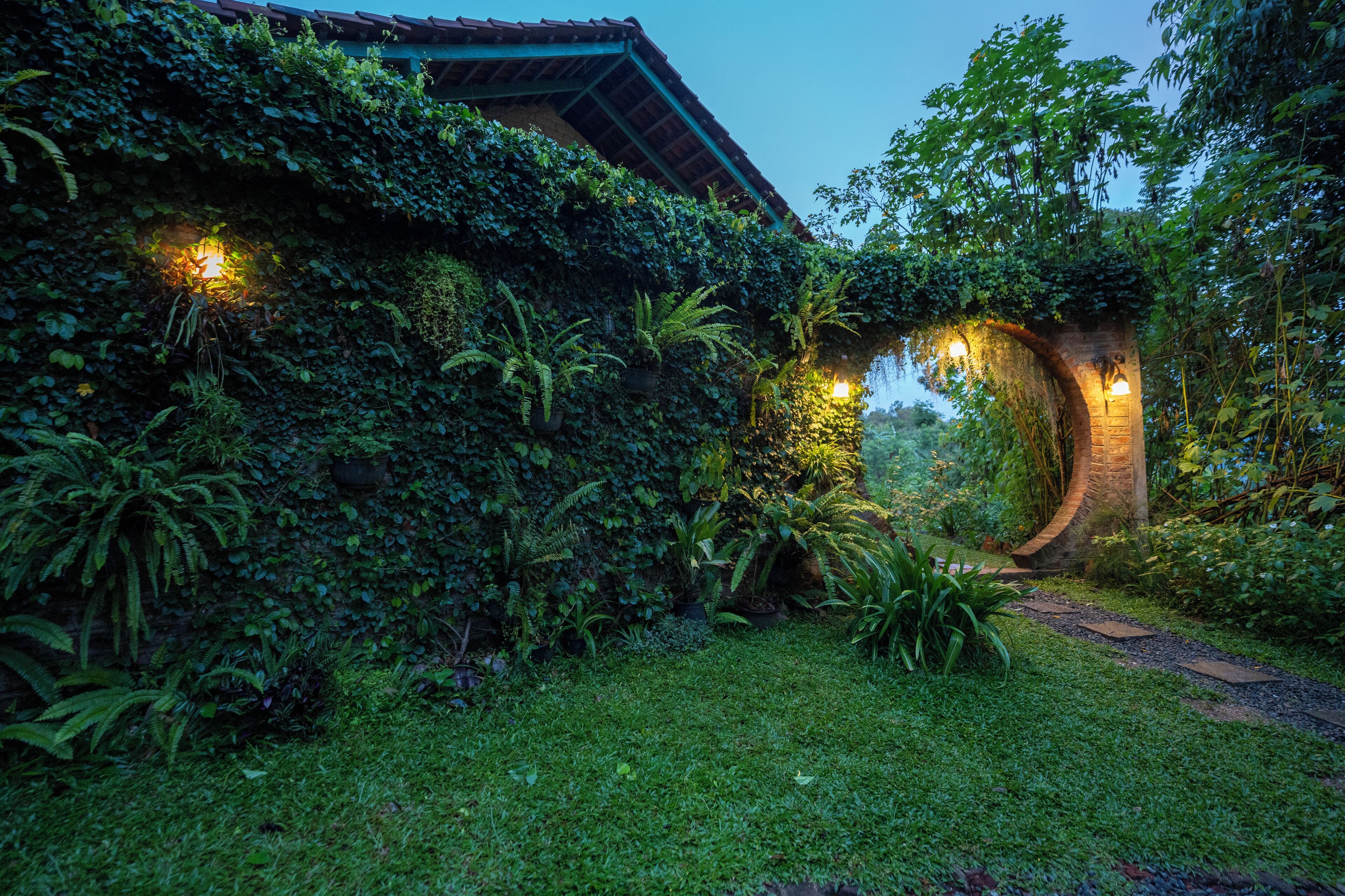 Un jardín exuberante con un arco de ladrillo, un sendero y un edificio parcialmente cubierto por follaje.