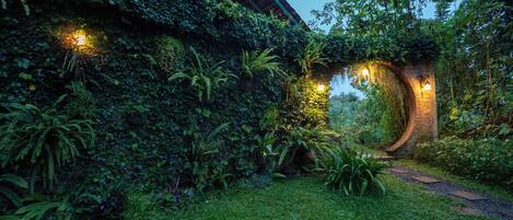 Un jardín exuberante con un arco de ladrillo, un sendero y un edificio parcialmente cubierto por follaje.
