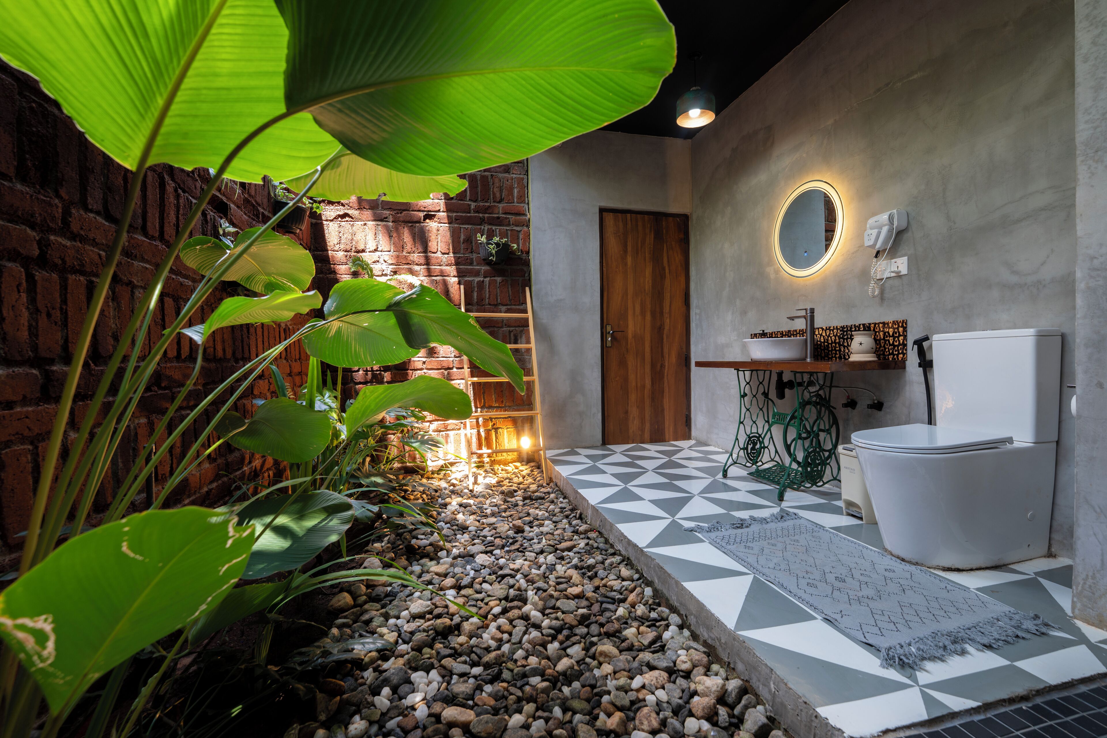 A bathroom with a tiled floor, a wooden door, a toilet, a sink, and a mirror with a light.