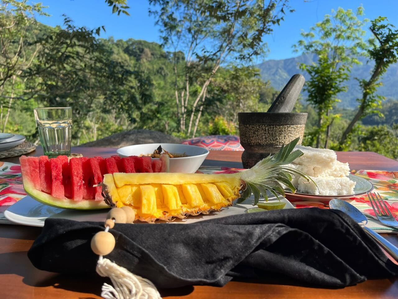 A table with a watermelon, pineapple, and rice, set outdoors with a mountain view.