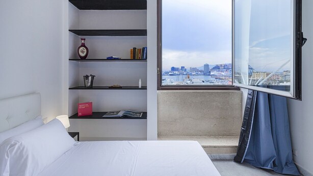 A hotel room with a bed, a bookshelf, and a window with a city view. - Chic Modern Lofts - B - BK (Genoa)
