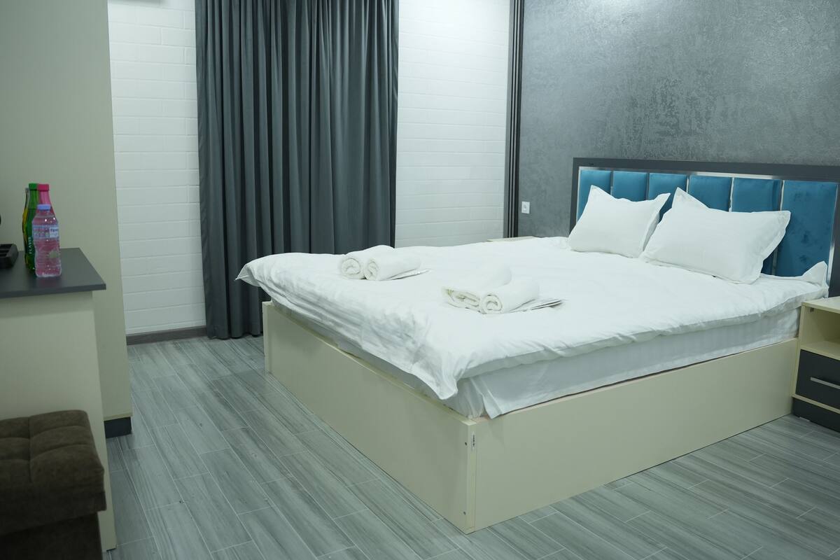 A neatly made bed with white linens and towels, a bedside table with a lamp, and a grey curtain.