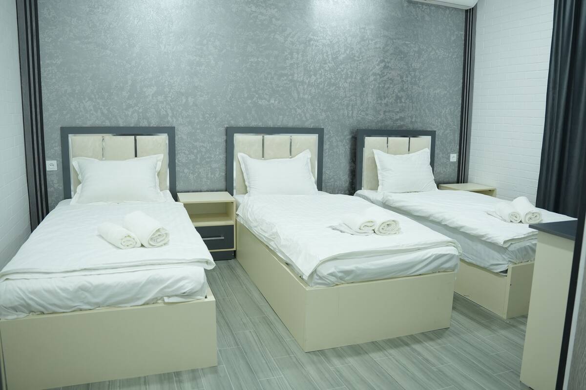 A hotel room with three single beds, each with a white mattress and pillow, and a small bedside table. The room has a grey textured wall and a white brick accent wall.