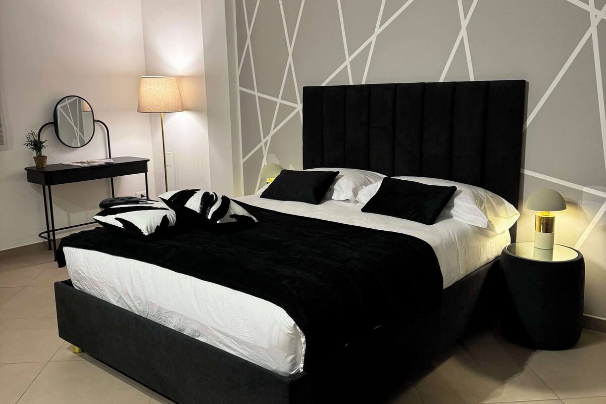 Une chambre moderne avec un lit noir, du linge de lit blanc, un motif géométrique sur le mur, une table de chevet avec une lampe et une petite plante.