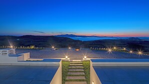 A modern outdoor staircase leading to a terrace with a view of a sunset over a body of water and hills.