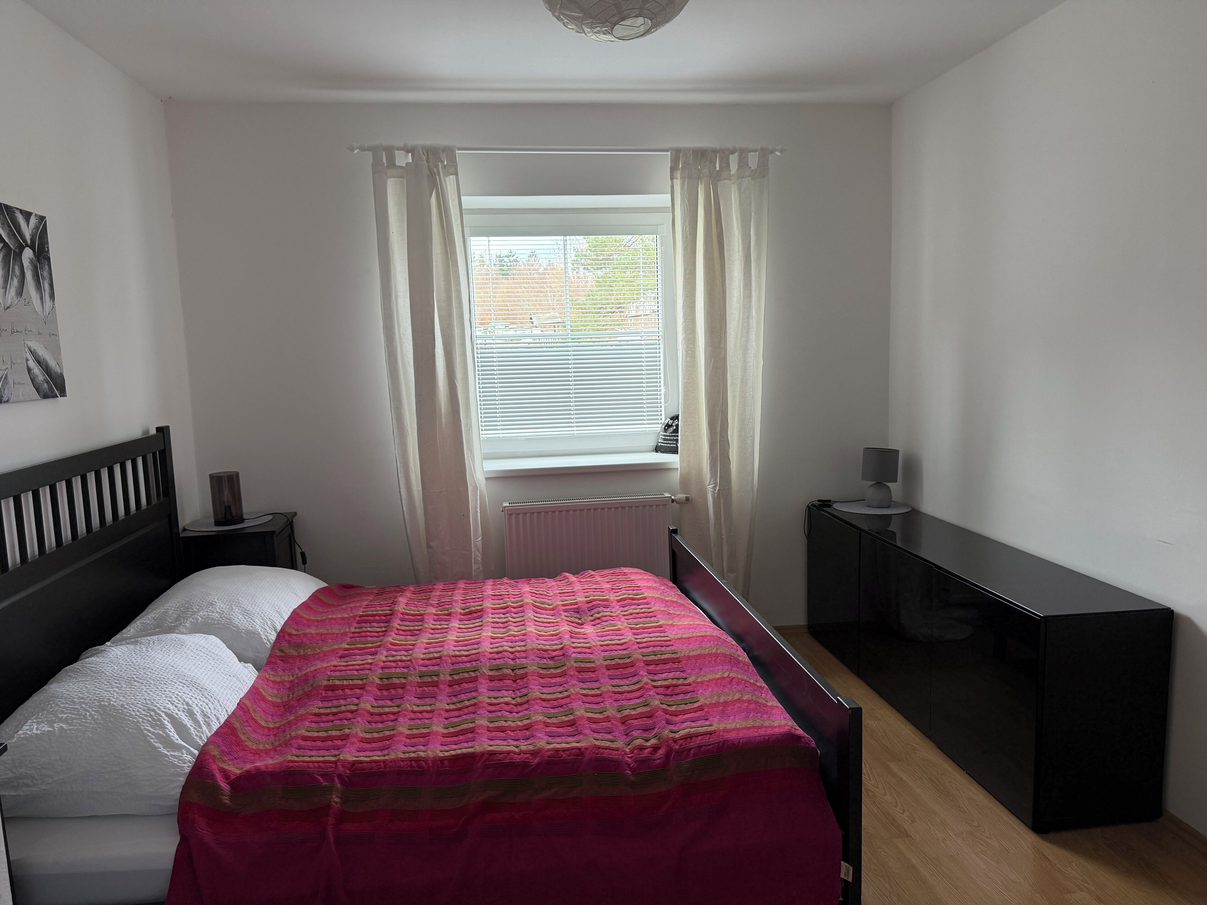 Quarto com cama, escrivaninha, radiador e janela com cortinas.