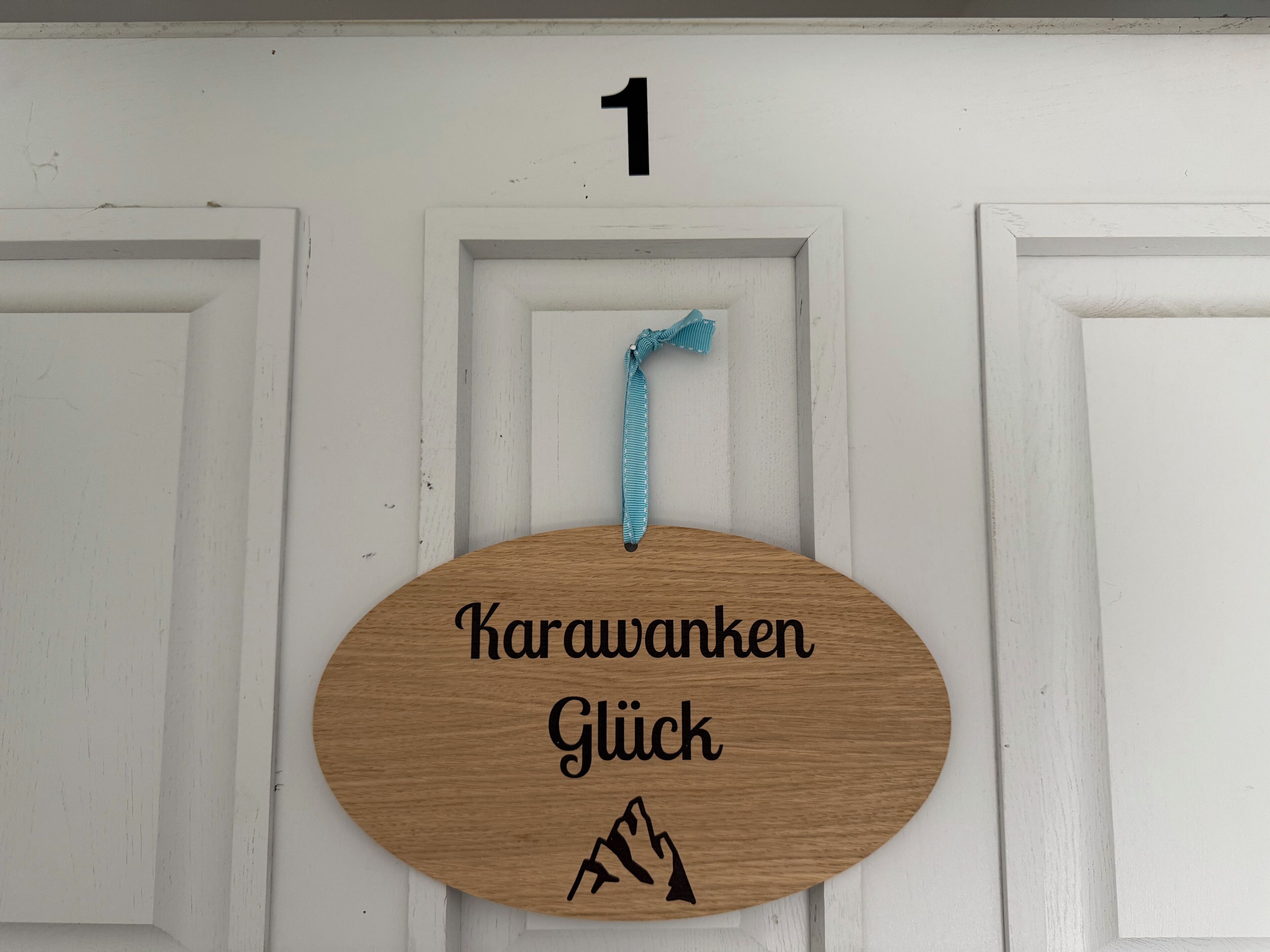 A wooden sign with the text "Karawanken Glück" and a mountain icon, hanging on a white door with the number 1 above it.