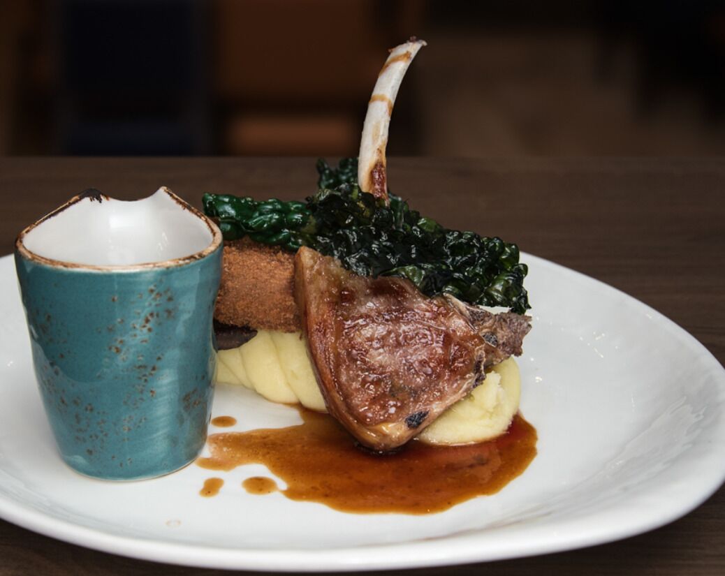 A plate with a lamb chop, mashed potatoes, and greens, accompanied by a blue ceramic cup.