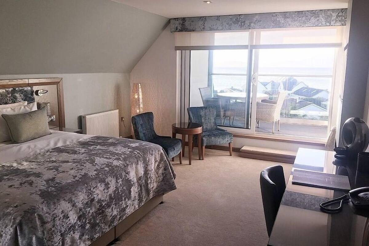 A hotel room with a large bed, a desk, two chairs, and a view of the sea through a large window.