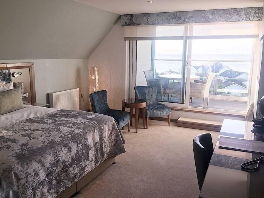 Habitación de hotel con una cama grande, un escritorio, dos sillas y vista al mar a través de un ventanal amplio.