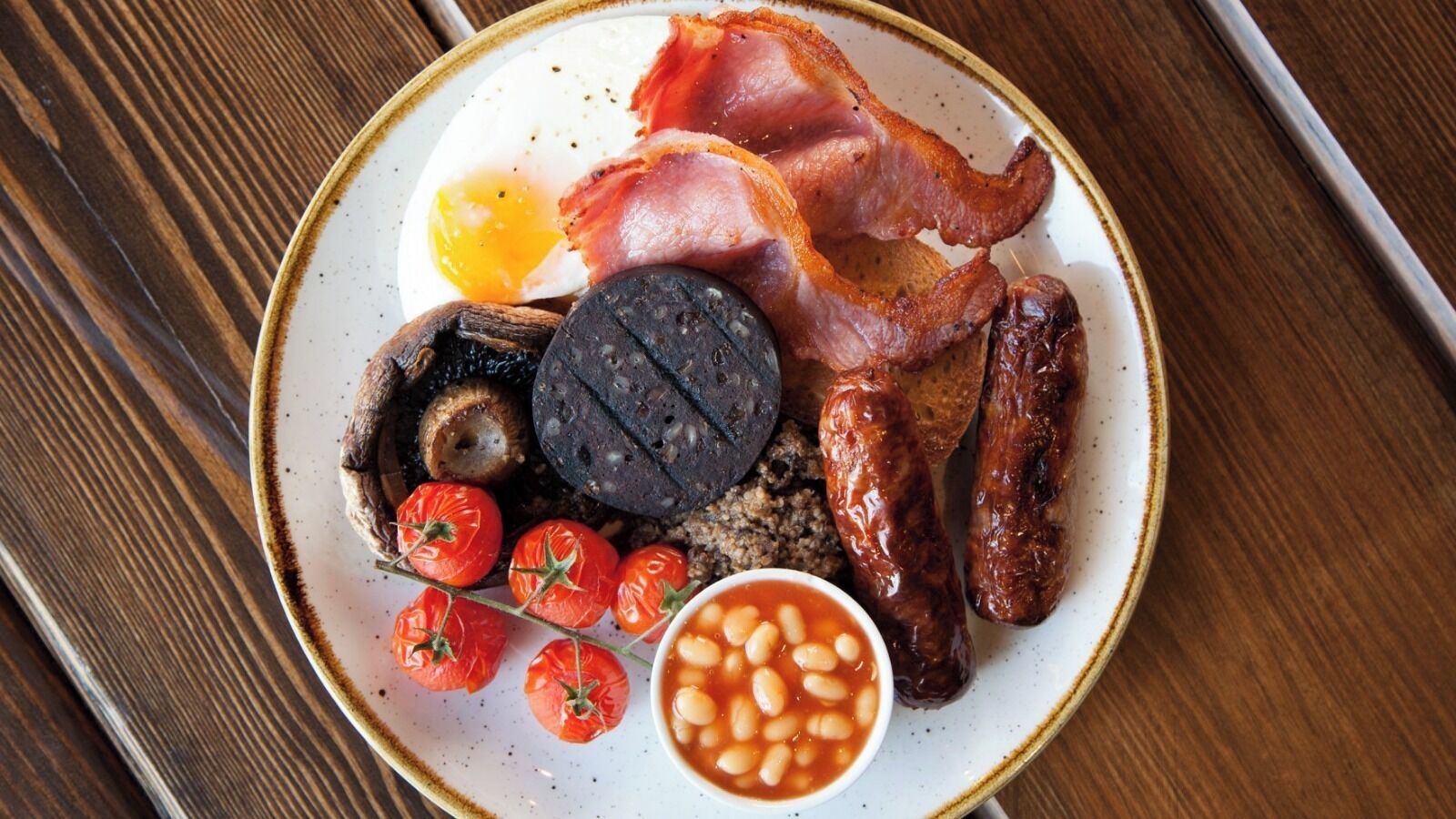 A plate of breakfast food including bacon, sausages, eggs, tomatoes, and beans.