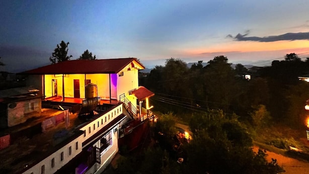 A two-story building with a red roof, illuminated interior, and a balcony. - Uday Homestay (Baijnath)